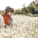 Side view of positive kid with outstretched arms in agricultural meadow with dried plants and green trees on blurred background